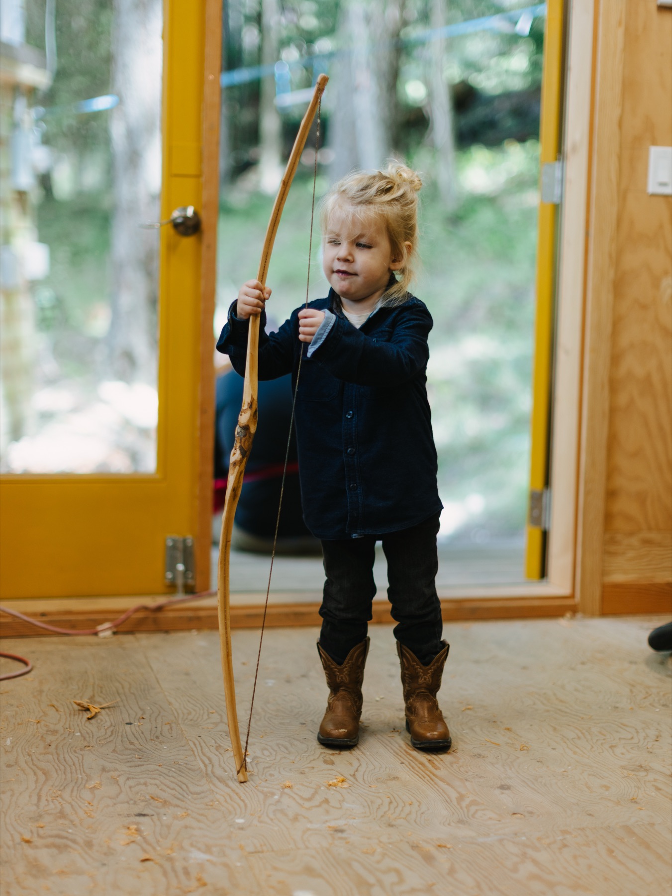 A young archer holding a finished wooden bow in the workshop doorway.