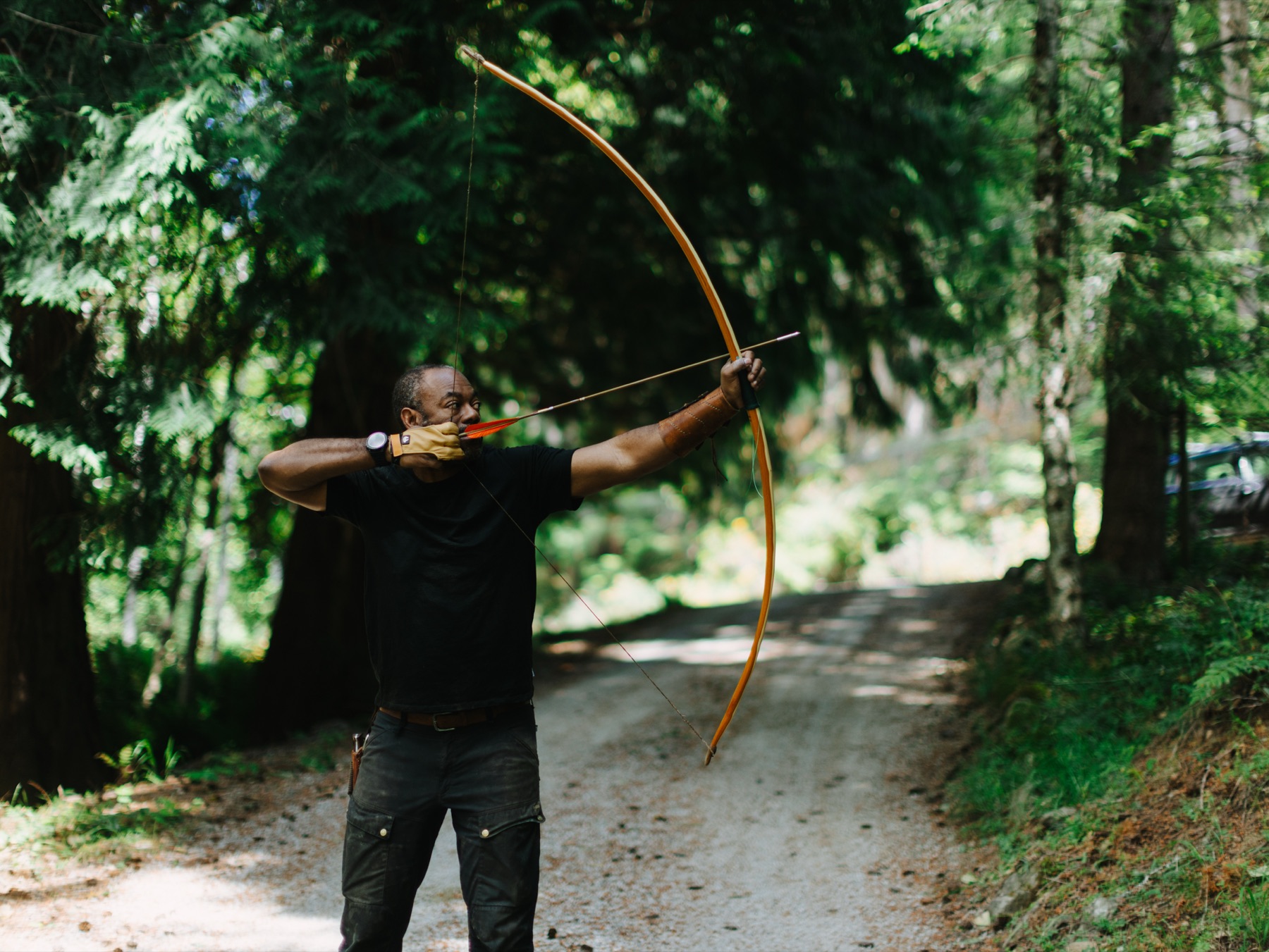 An archer at full draw with a wooden longbow on a forest road.